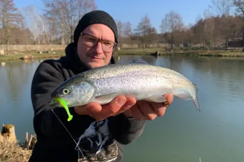 Glücklicher Angler hält frisch gefangene Forelle am Forellenteich. Der Fisch hat einen grellgrünen Köder im Maul.