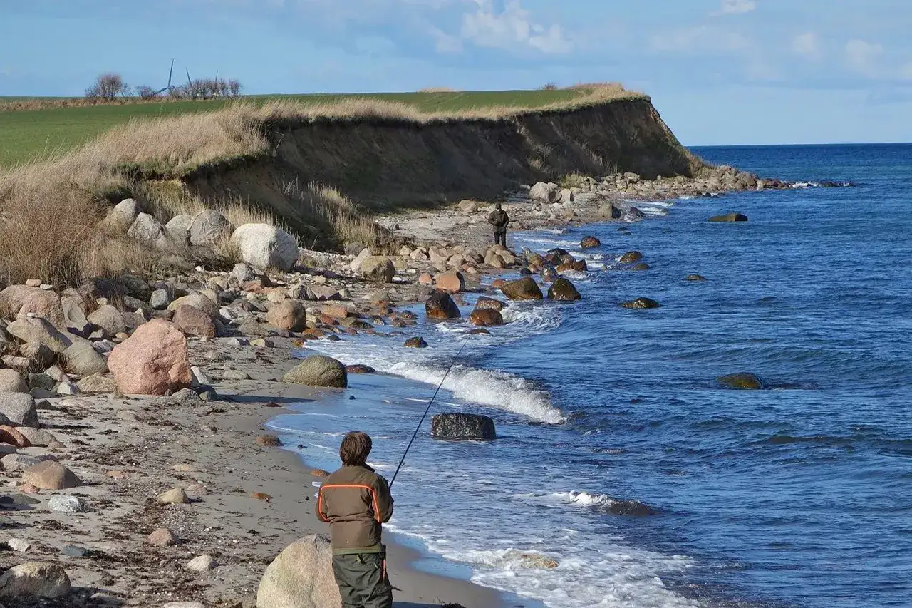 Ein Angler steht am steinigen Strand von Langeland und wirft seine Angel aus. Die Steilküste im Hintergrund und das blaue Meer sind aktuell ein idyllisches Bild.